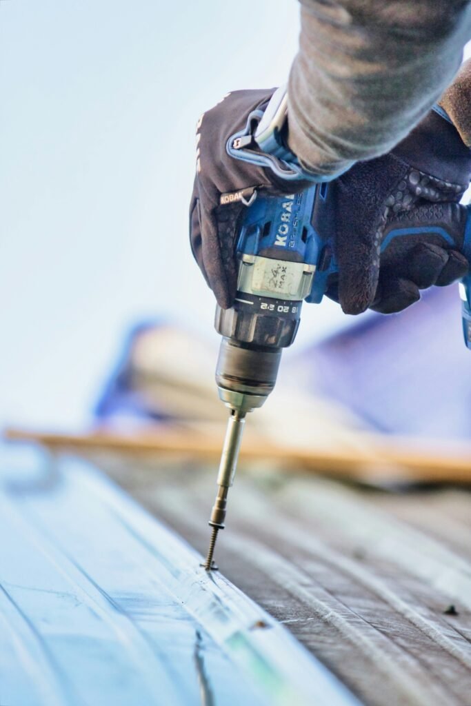 Close-up of a worker’s gloved hand using a Kobalt power drill to fasten a screw into a metal surface, with a blurred background.
