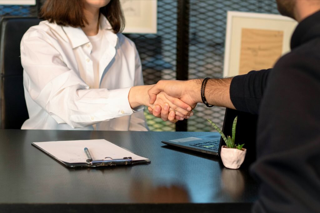 Two people shaking hands across a desk during a professional meeting, with a clipboard, pen, and laptop visible on the table.