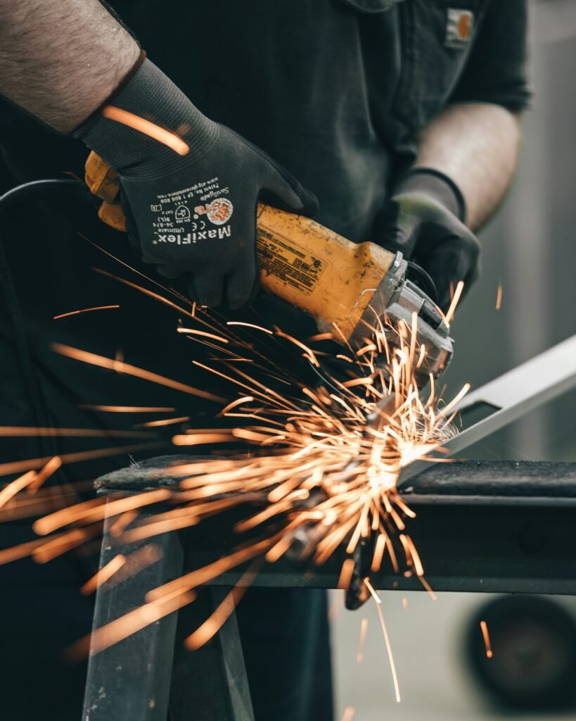 Man wearing protective gloves and welding helmet using an angle grinder on metal, emitting bright sparks in an industrial workshop.